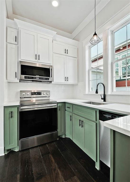 A modern kitchen corner with white upper cabinets, sage green lower cabinets, stainless steel appliances, dark wood floors, and a black faucet under a window.