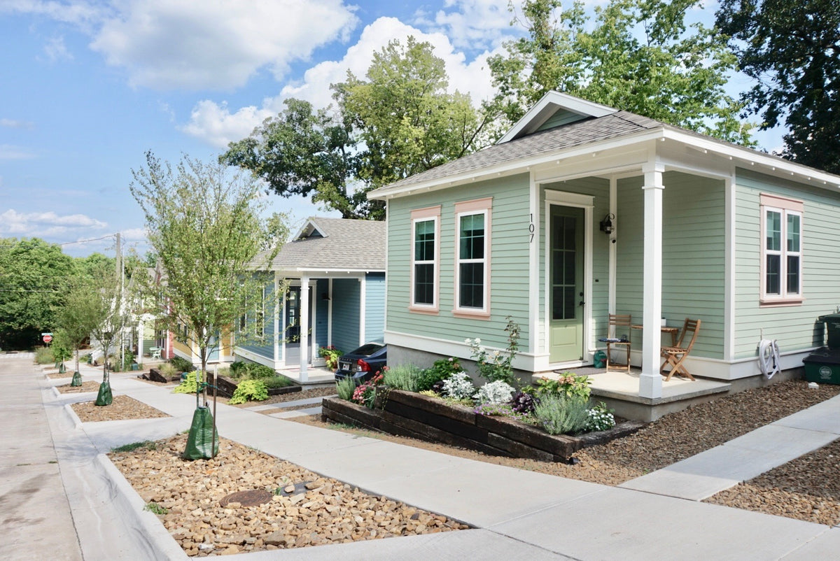 A light sage green home with white trim and a front porch, shown on a sunny day with trees, sky, and neighboring houses in the background.