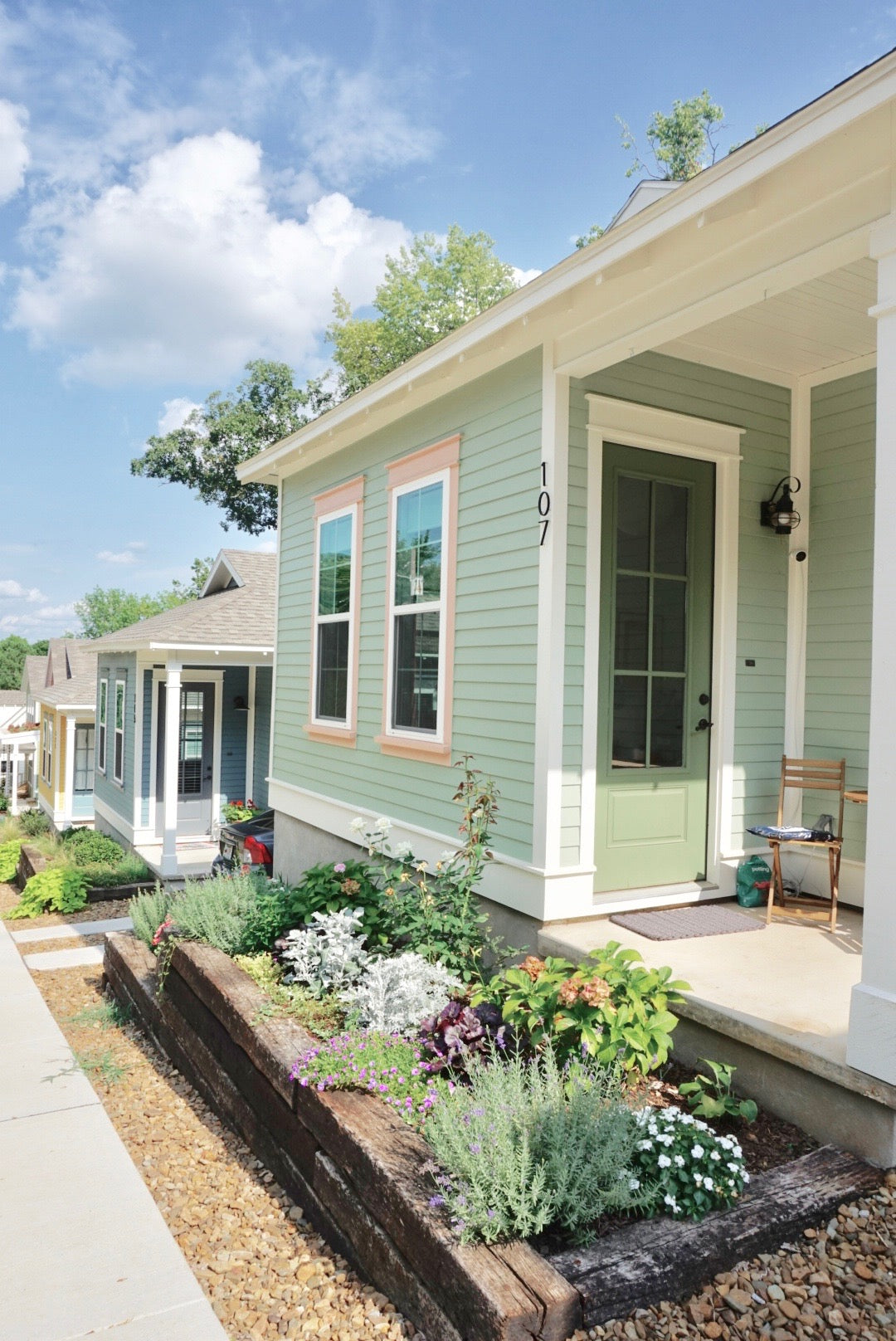 A light sage green cottage-style house with white trim, a front porch, and a landscaped flower bed in the foreground under a blue sky.