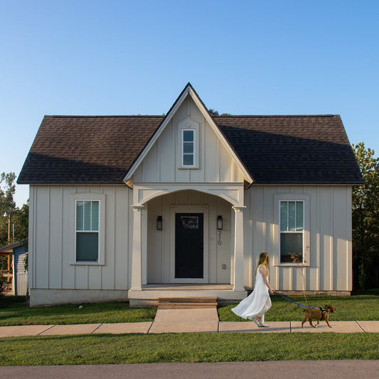 Woman in a white dress walking a dog in front of a small house on a sunny day.