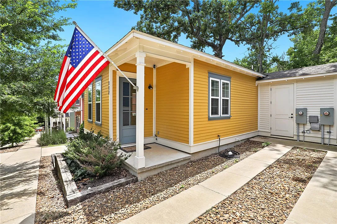 A bright yellow single-story cottage with white trim, a front porch, an American flag on the porchpost, a gravel walkway,