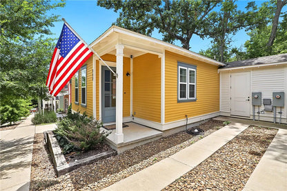 A bright yellow single-story cottage with white trim, a front porch, an American flag on the porchpost, a gravel walkway,